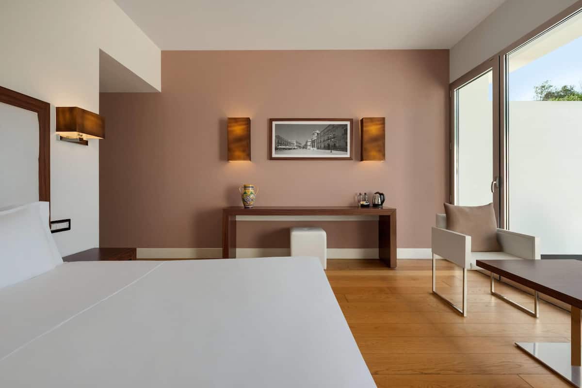 Ein modernes Hotelzimmer mit einem großen Bett, einem Schreibtisch mit Stuhl, einer Kaffeemaschine und einem gerahmten Bild an der Wand.