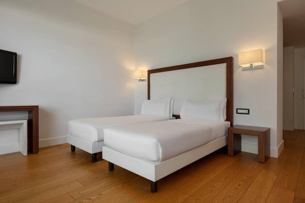 Ein Doppelbett mit weißer Bettwäsche, zwei Nachttische, ein an der Wand befestigter Fernseher und ein kleiner Holztisch.