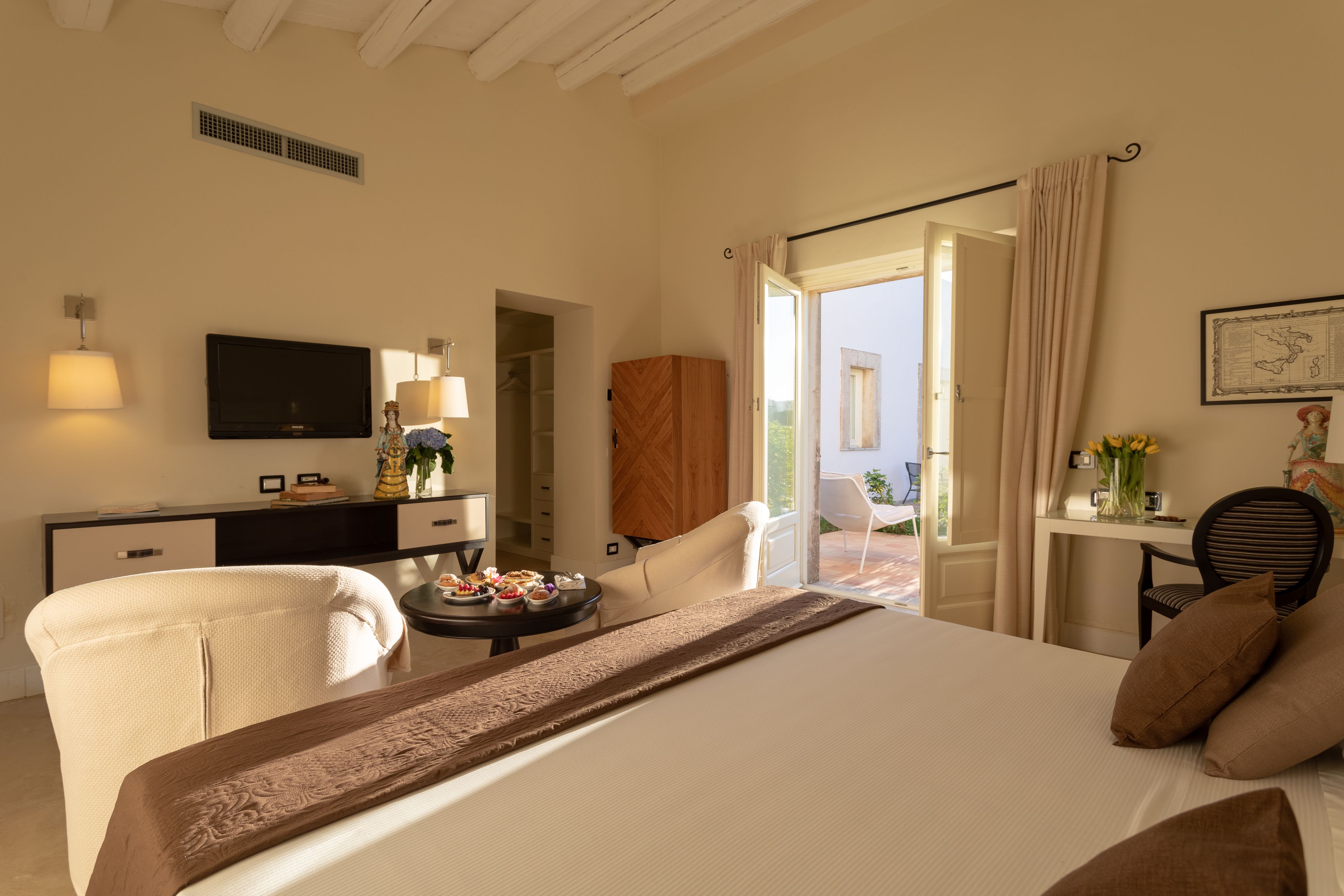 A hotel room with a bed, a desk with a flat-screen TV, a chair, a small table with food, and a view of the outdoors.