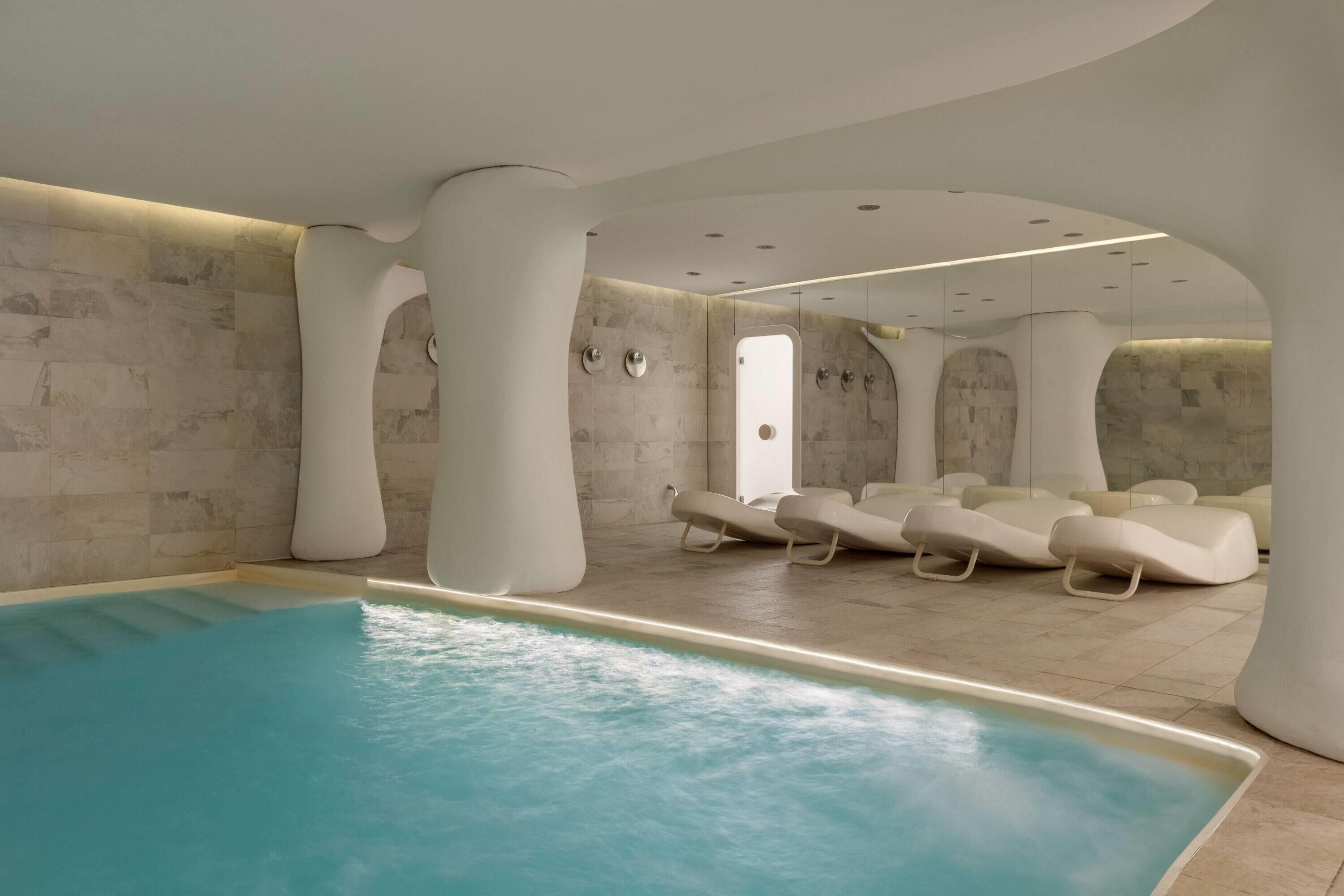 Una piscina interior moderna con una superficie de agua azul cristalina, rodeada de sillones blancos y un diseño arquitectónico único con pilares curvos.