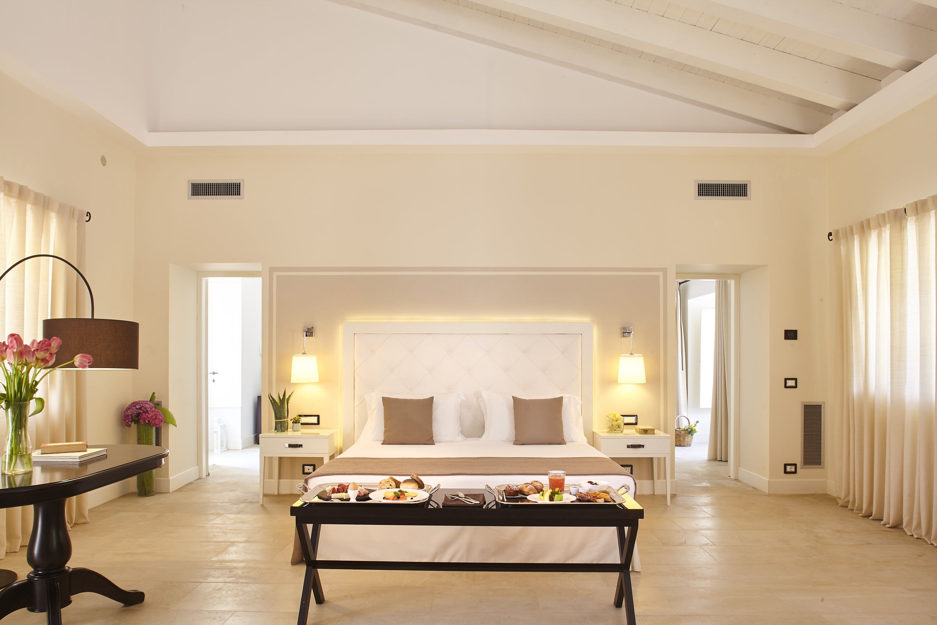 A spacious hotel room with a large bed, bedside tables, a dining table with breakfast, and a view of the hallway.