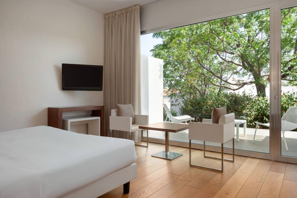 Ein modernes Hotelzimmer mit einem großen Bett, einem an der Wand befestigten Fernseher, einer Essecke mit Stühlen und einem Tisch sowie einem Balkon mit Blick auf Bäume und Gartenmöbel.