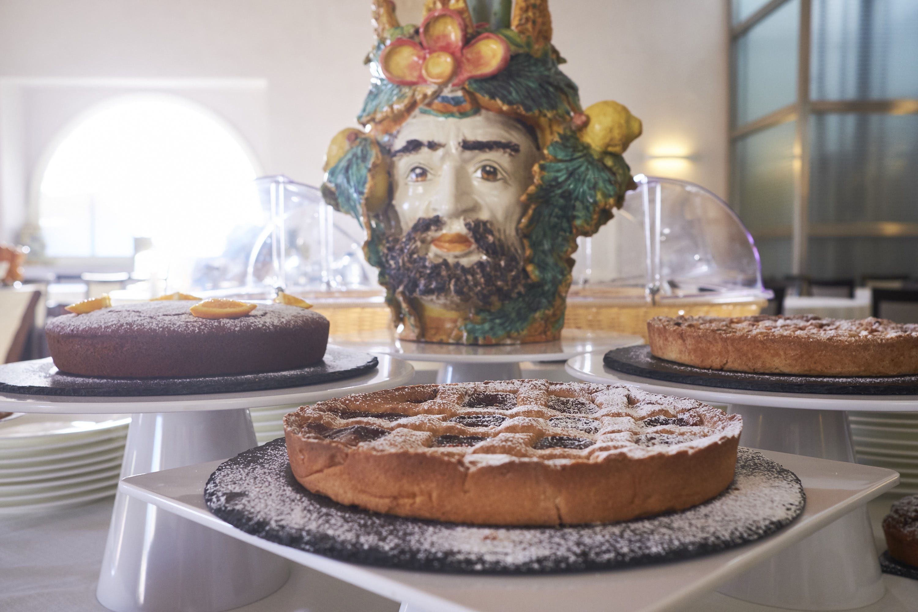 A dessert display with various cakes on white stands, a decorative ceramic piece, and a glass container in the background.