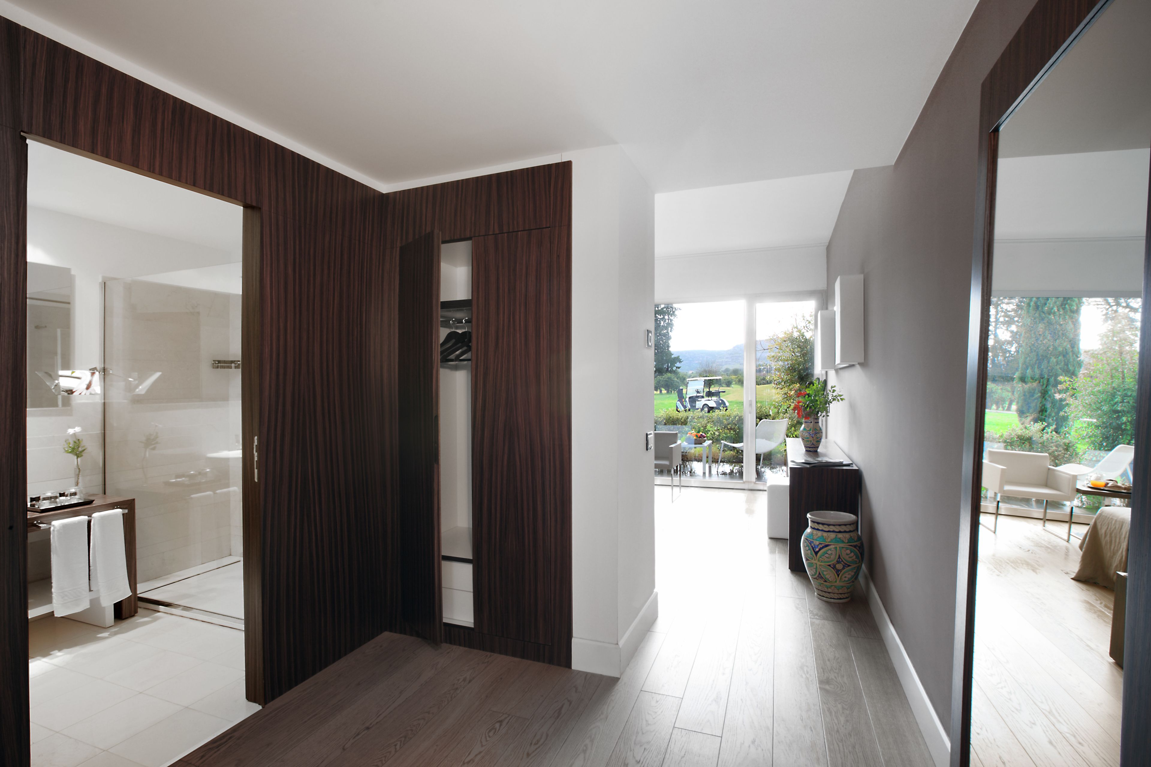 A modern interior with a wooden floor, a glass door leading to a bathroom, and a view of a patio with outdoor furniture.