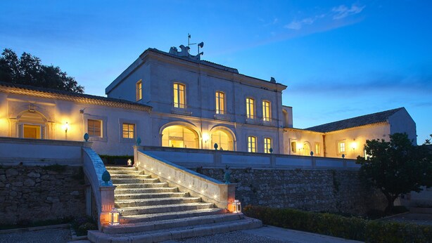 A large, multi-story building with a grand staircase leading to the entrance, stone walls, and multiple illuminated windows. - Dolce by Wyndham Siracusa I Monasteri Golf & Spa (Syracuse)
