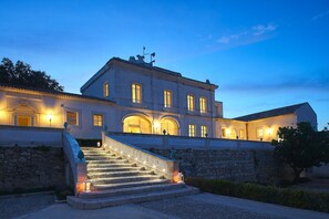 A large, multi-story building with a grand staircase leading to the entrance, stone walls, and multiple illuminated windows. - Dolce by Wyndham Siracusa I Monasteri Golf & Spa (Syracuse)