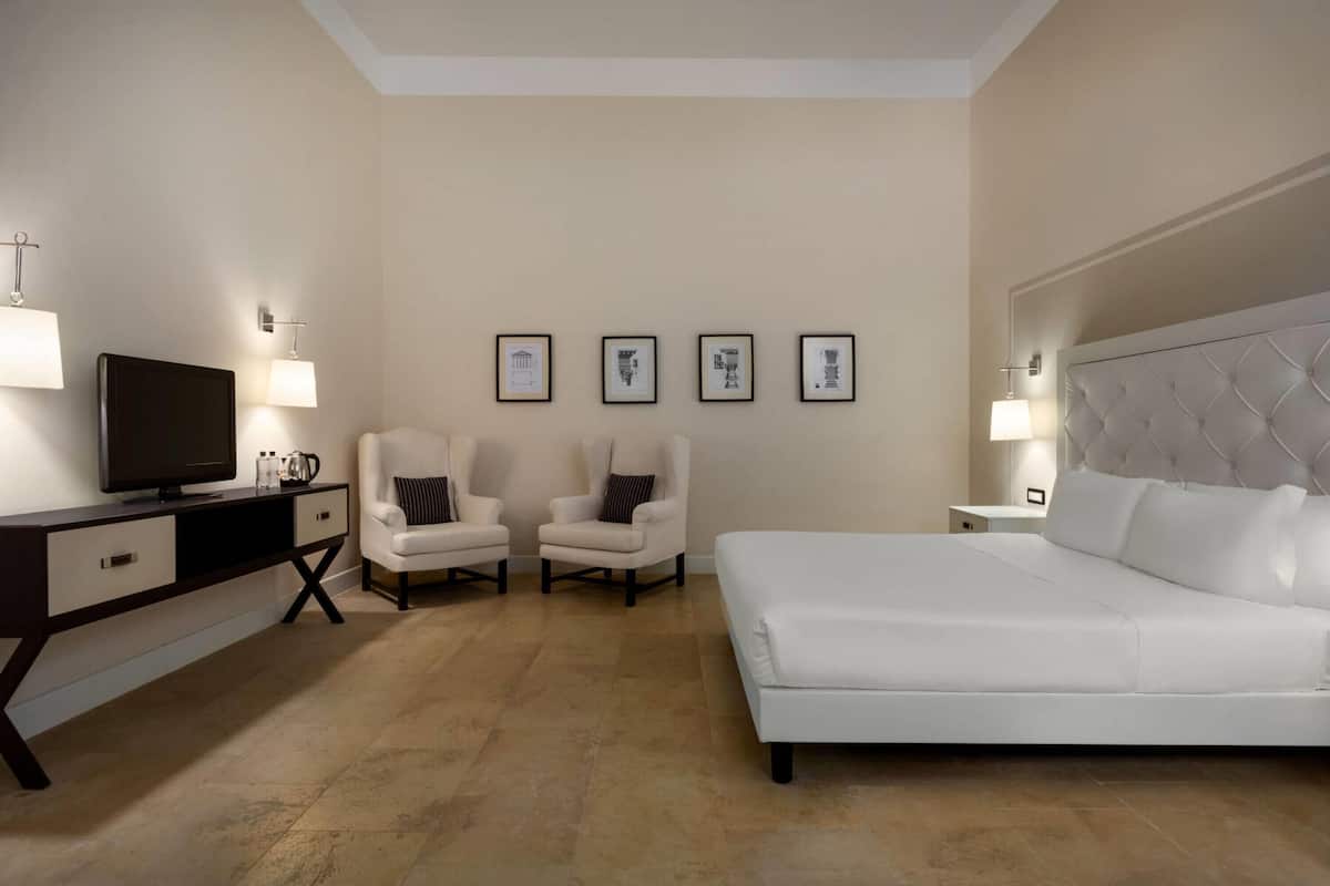 Ein Hotelzimmer mit einem Bett, zwei Sesseln, einem Schreibtisch mit Fernseher und gerahmten Bildern an der Wand.