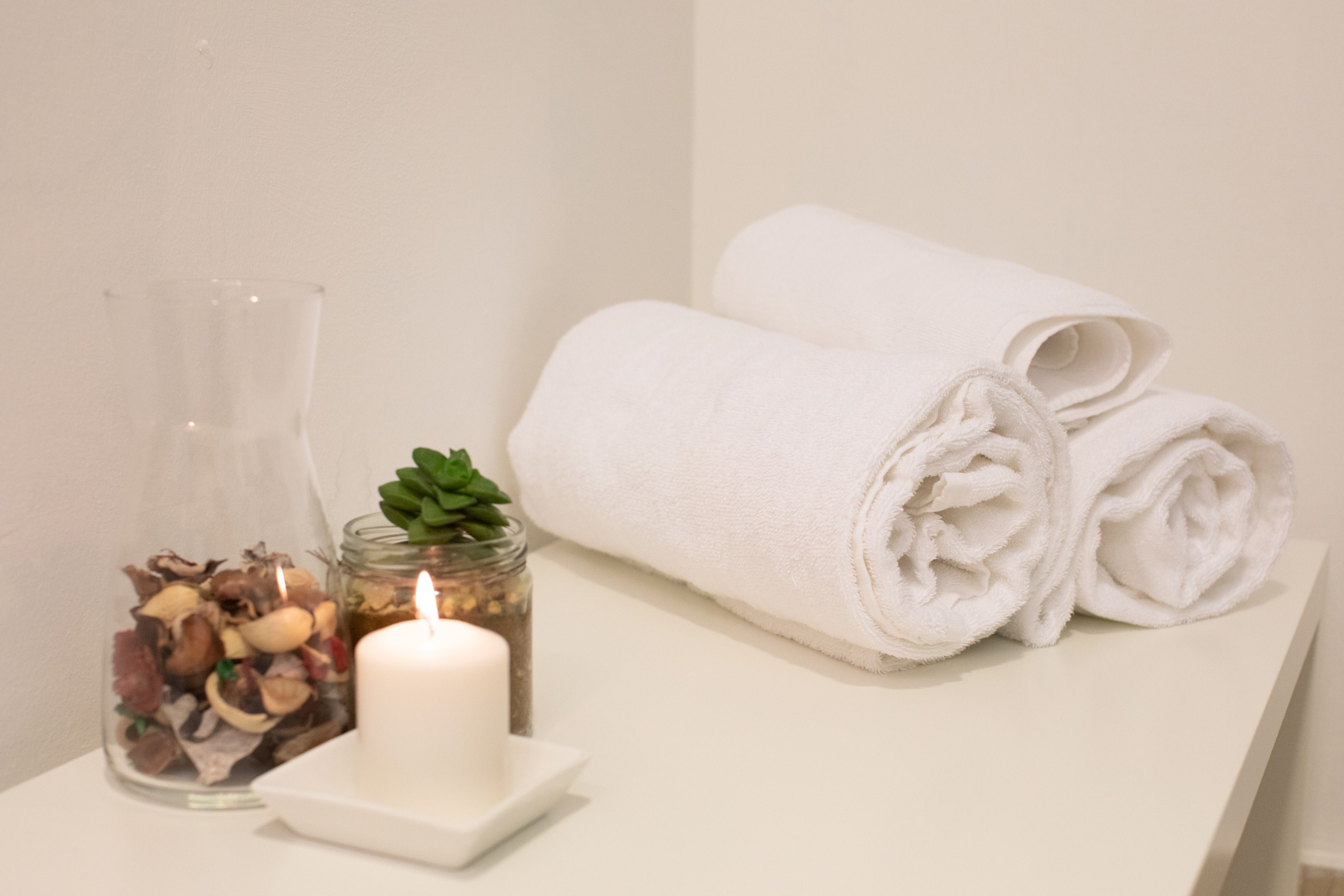 A white table with a glass jar containing dried mushrooms, a lit candle, and a rolled white towel.