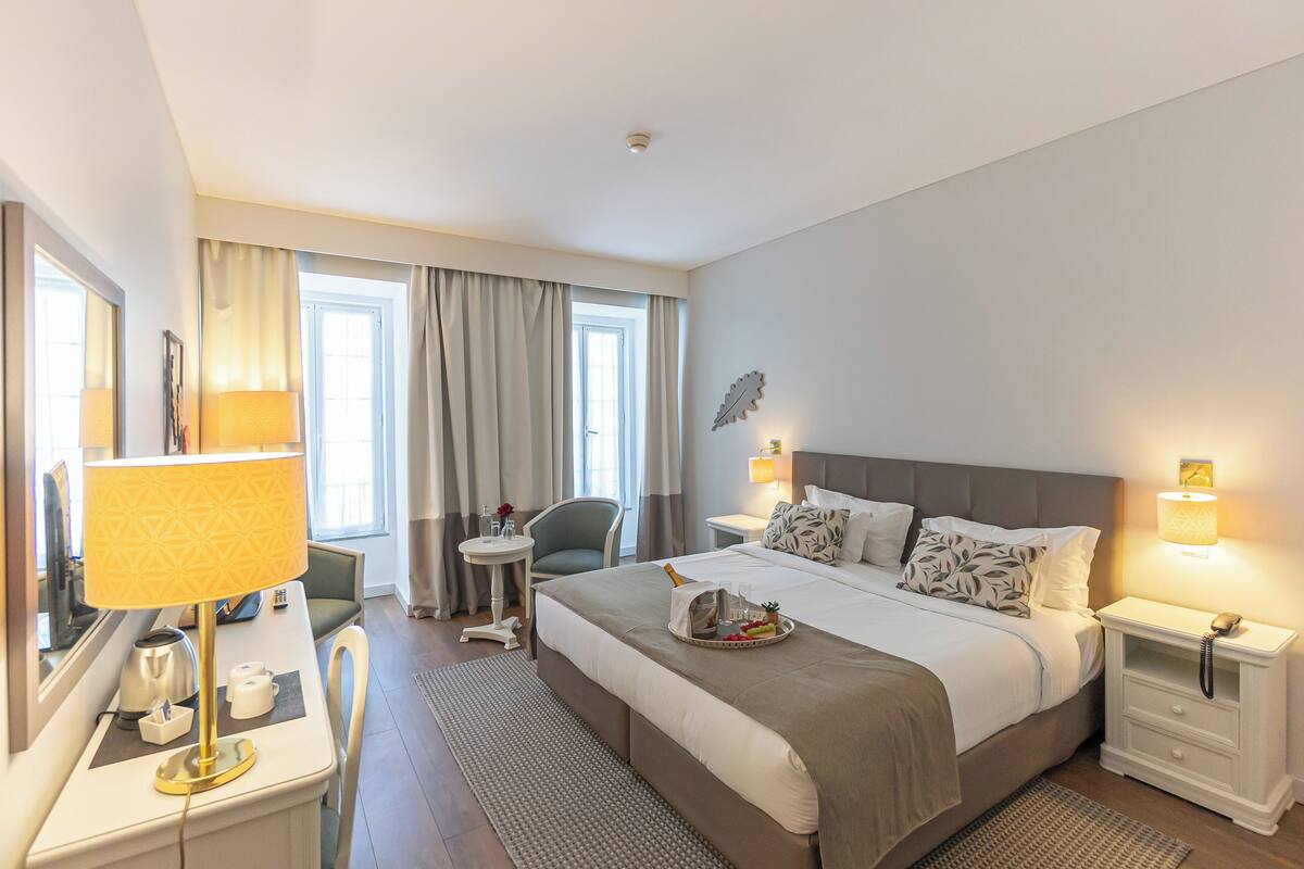 A hotel room with a large bed, bedside tables, a desk with a lamp, a chair, and a window with curtains.
