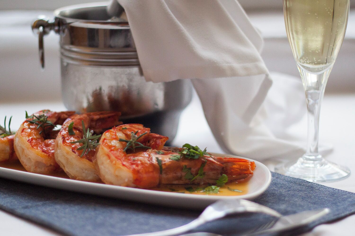 Une assiette de crevettes garnie d’herbes, un verre de vin blanc et un récipient en métal à l’arrière-plan.