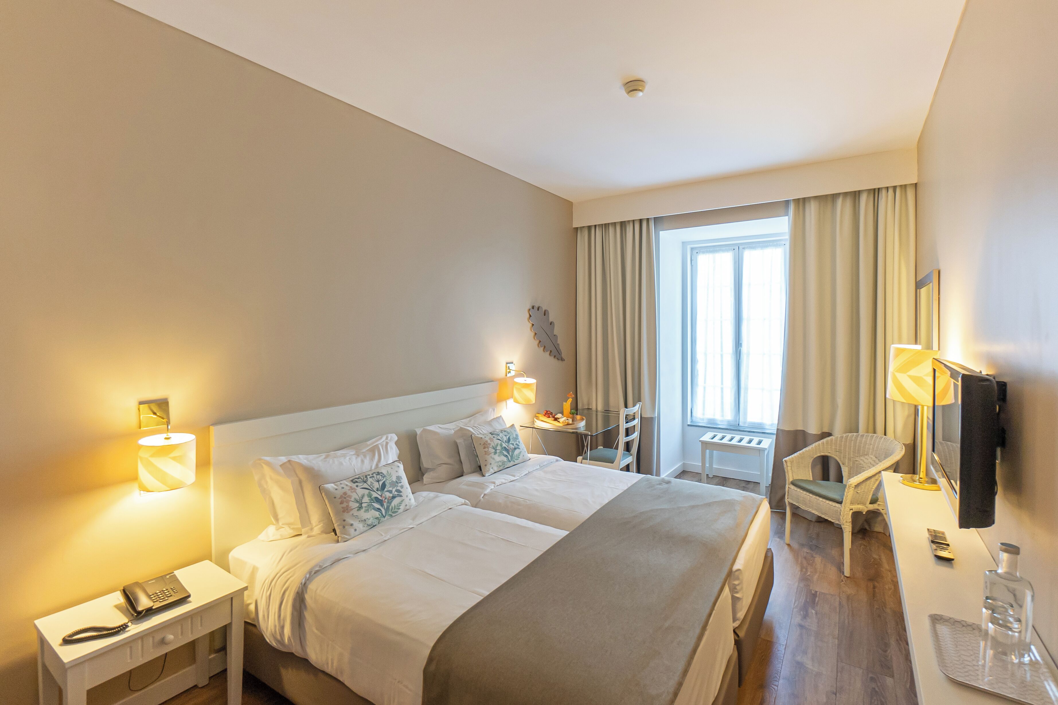 A hotel room with a large bed, bedside lamps, a desk with a chair, a TV, and a window with curtains.