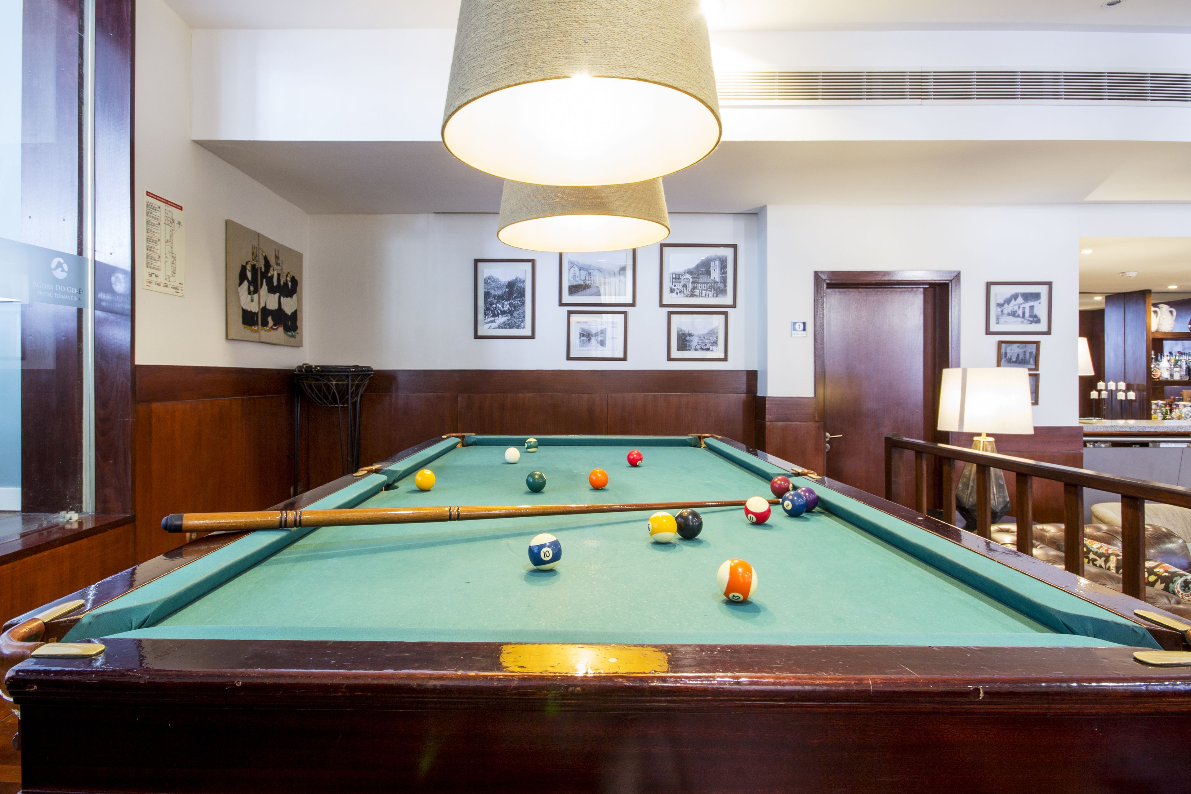 A pool table with balls, a pool cue, and a hanging lamp in a room with framed pictures on the wall.