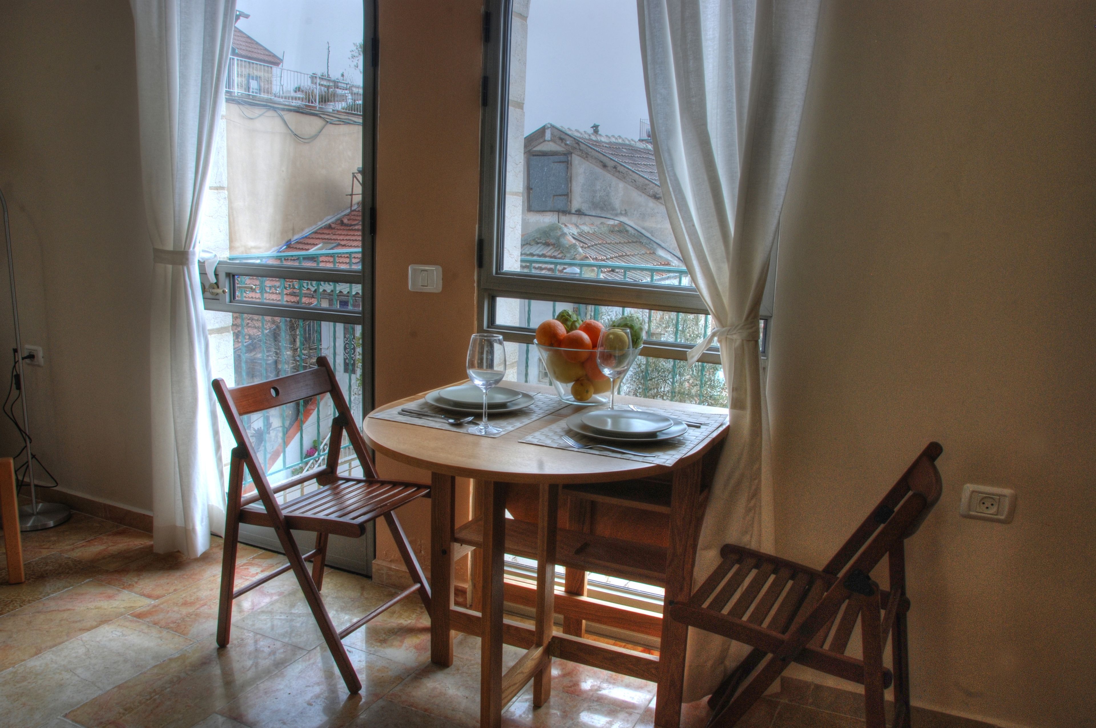 A dining area with a table set for two, a bowl of fruit, and a view of buildings through a window.