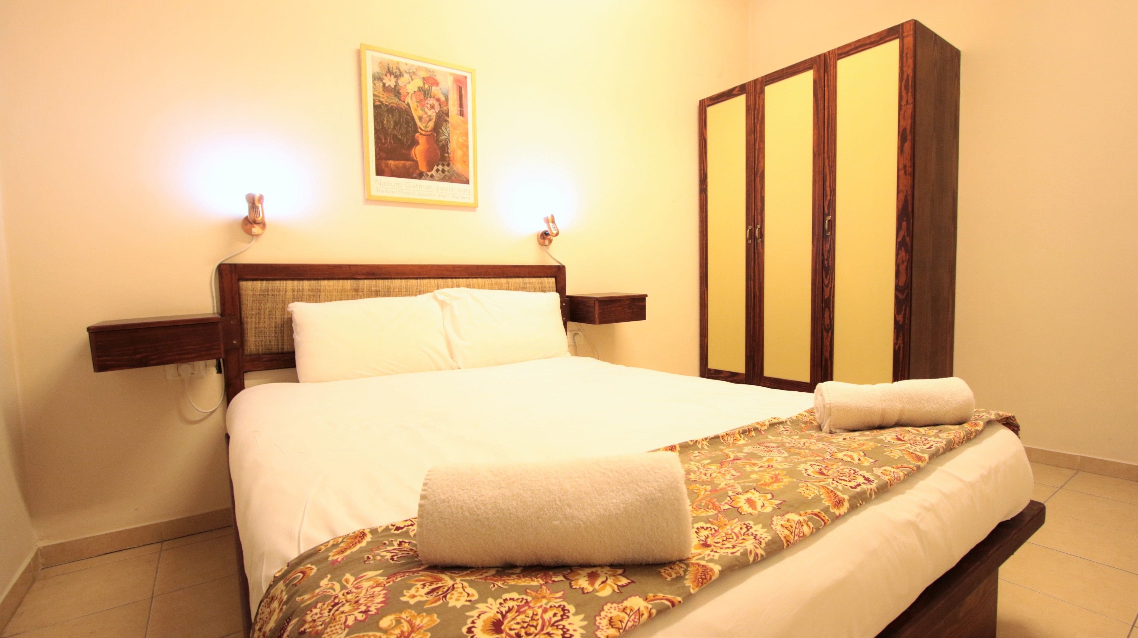 A neatly made bed with a floral bedspread and two pillows, a wooden nightstand with a lamp, a wardrobe with mirrored doors, and a framed artwork on the wall.