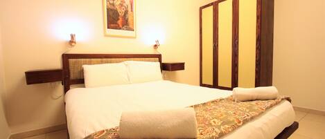 A neatly made bed with a floral bedspread and two pillows, a wooden nightstand with a lamp, a wardrobe with mirrored doors, and a framed artwork on the wall.