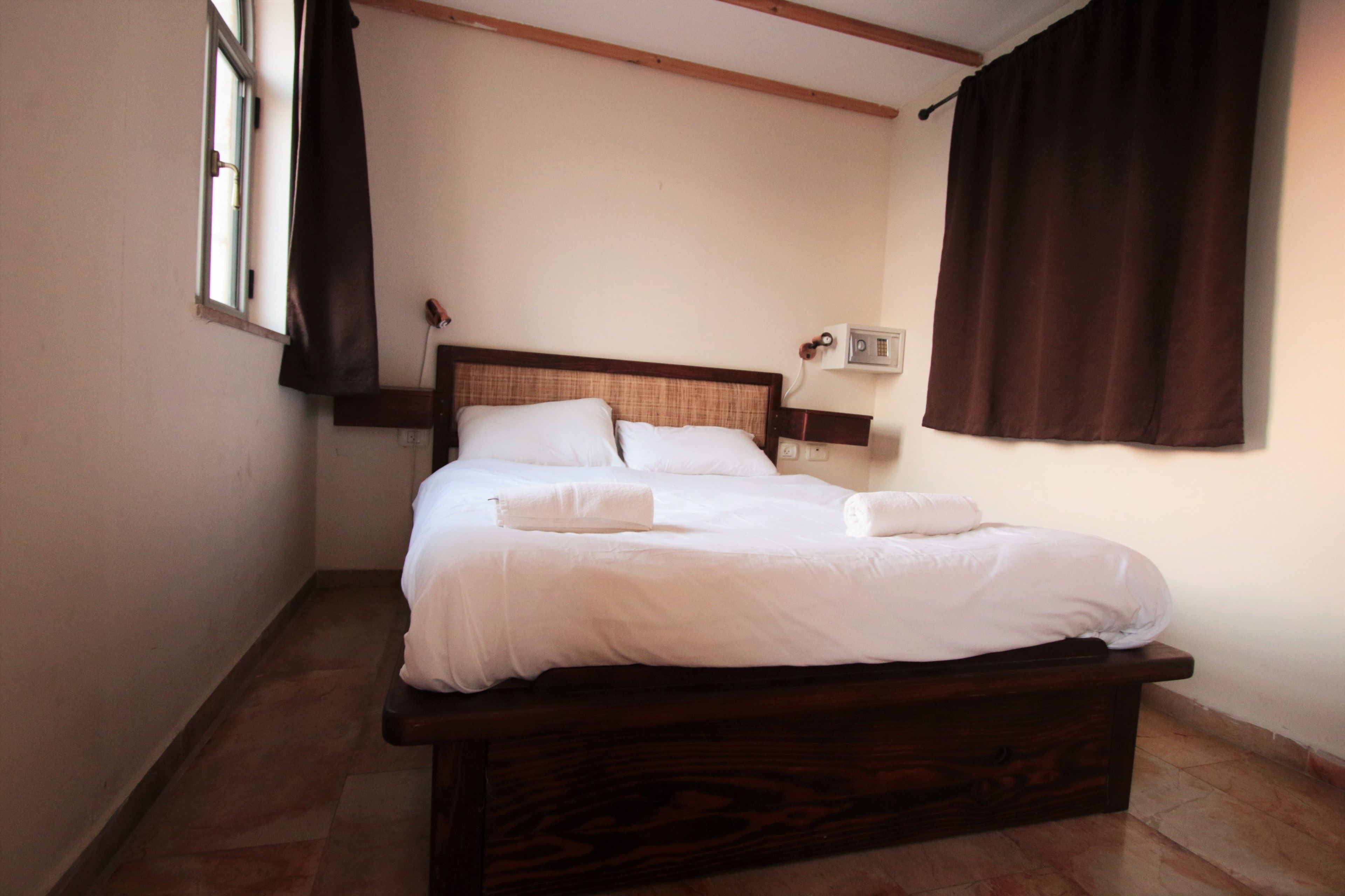 A neatly made bed with white linens and two pillows, a bedside table with a lamp, and a window with curtains.