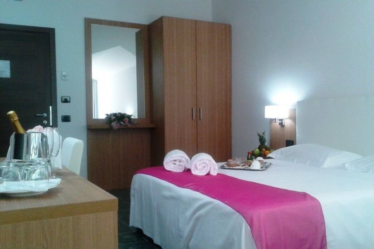 A hotel room with a bed, a desk with a bottle of champagne, a mirror, and a nightstand with a lamp.