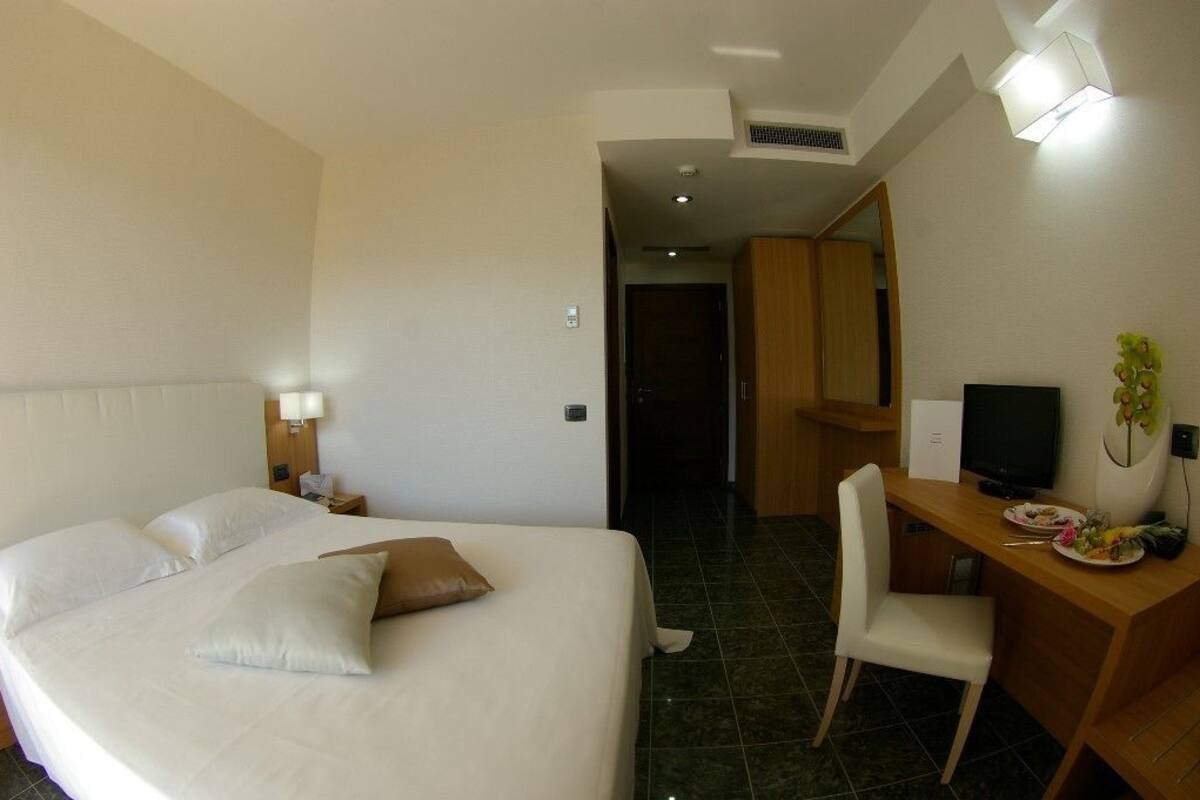 A hotel room with a large bed, a desk with a computer, a chair, and a small table with a plant and some items on it.