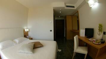 A hotel room with a large bed, a desk with a computer, a chair, and a small table with a plant and some items on it.