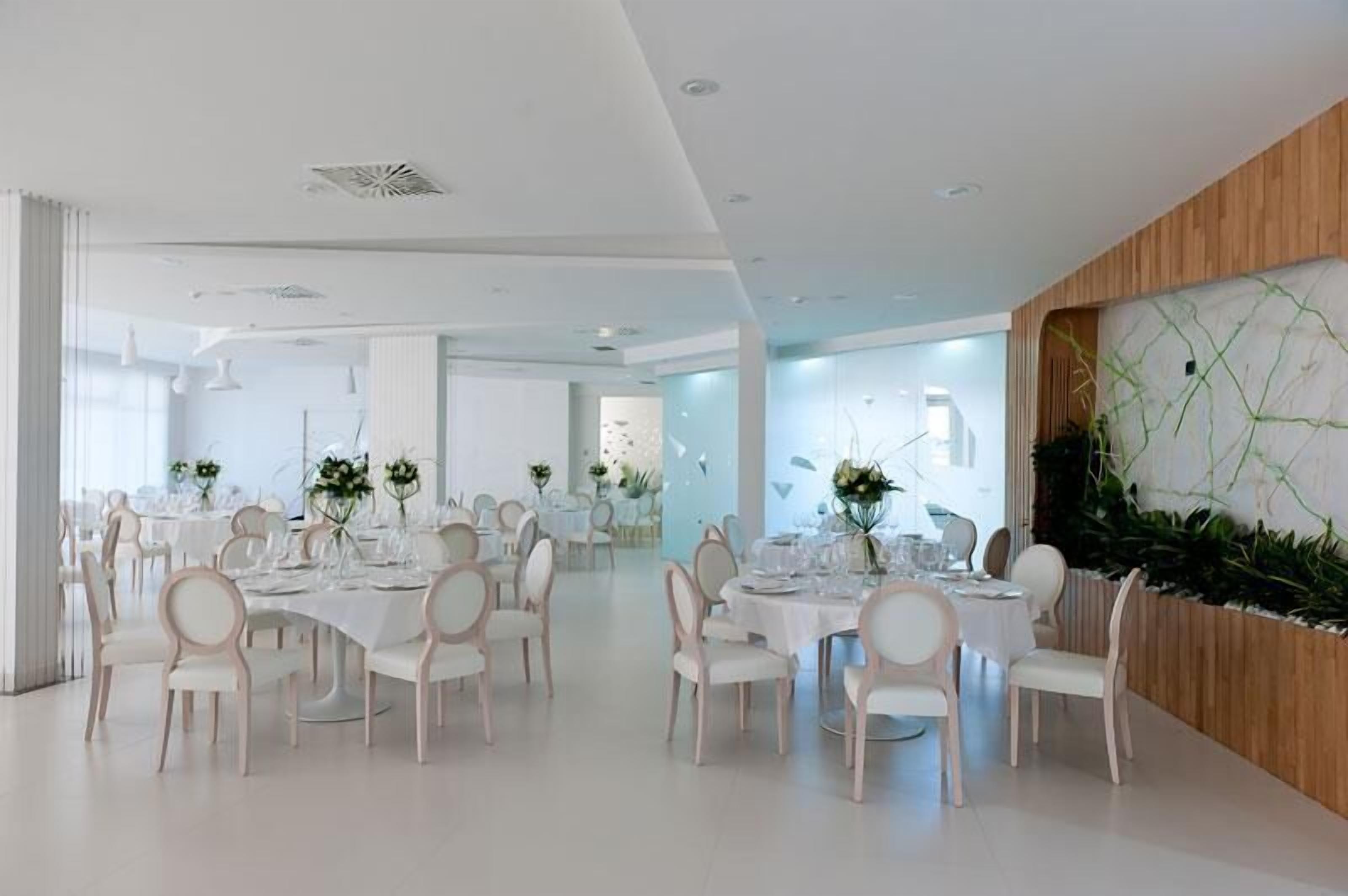 Un comedor moderno con mesas y sillas blancas, un panel de acento de madera y una planta grande.