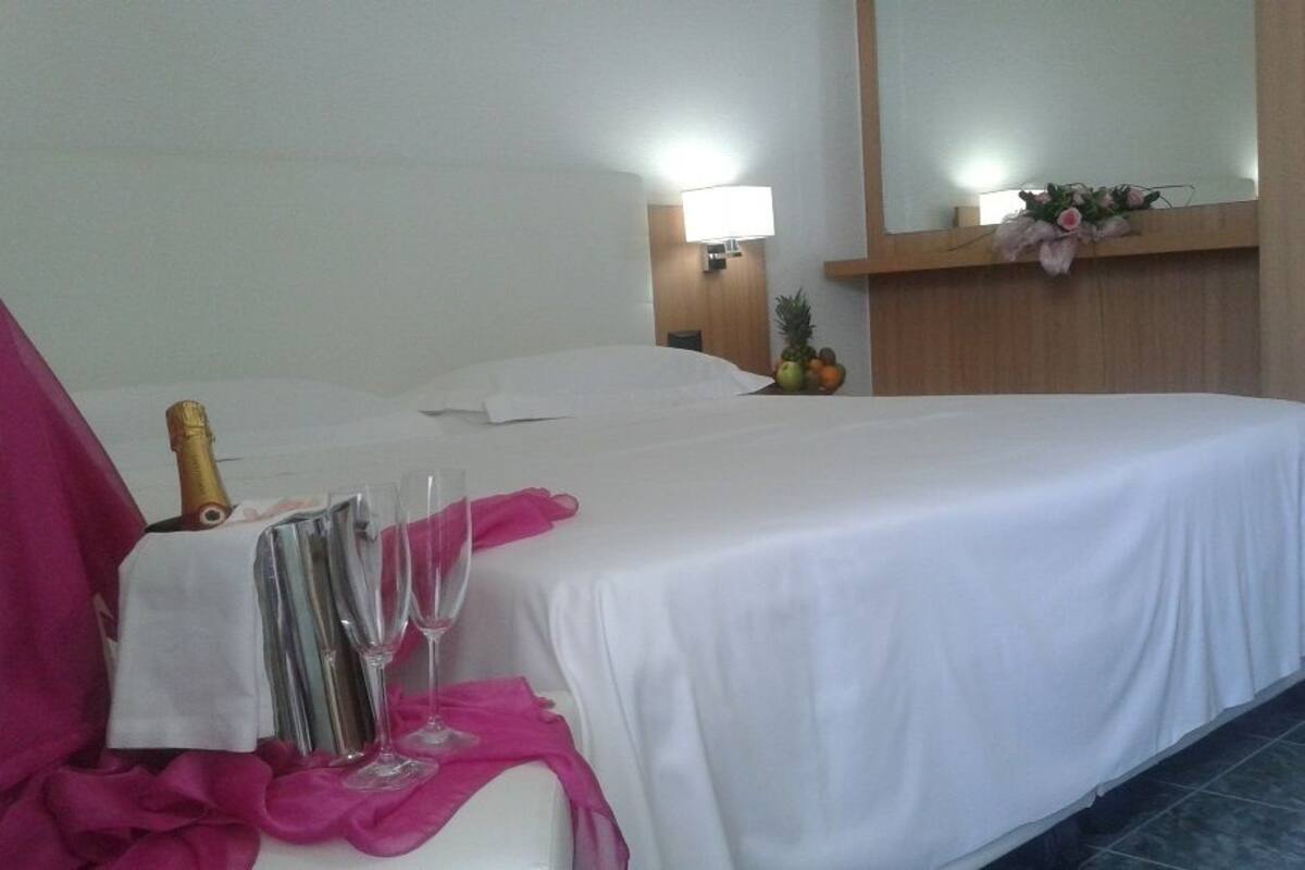A hotel room with a bed, a nightstand, a mirror, and a small table with a bottle and glasses.