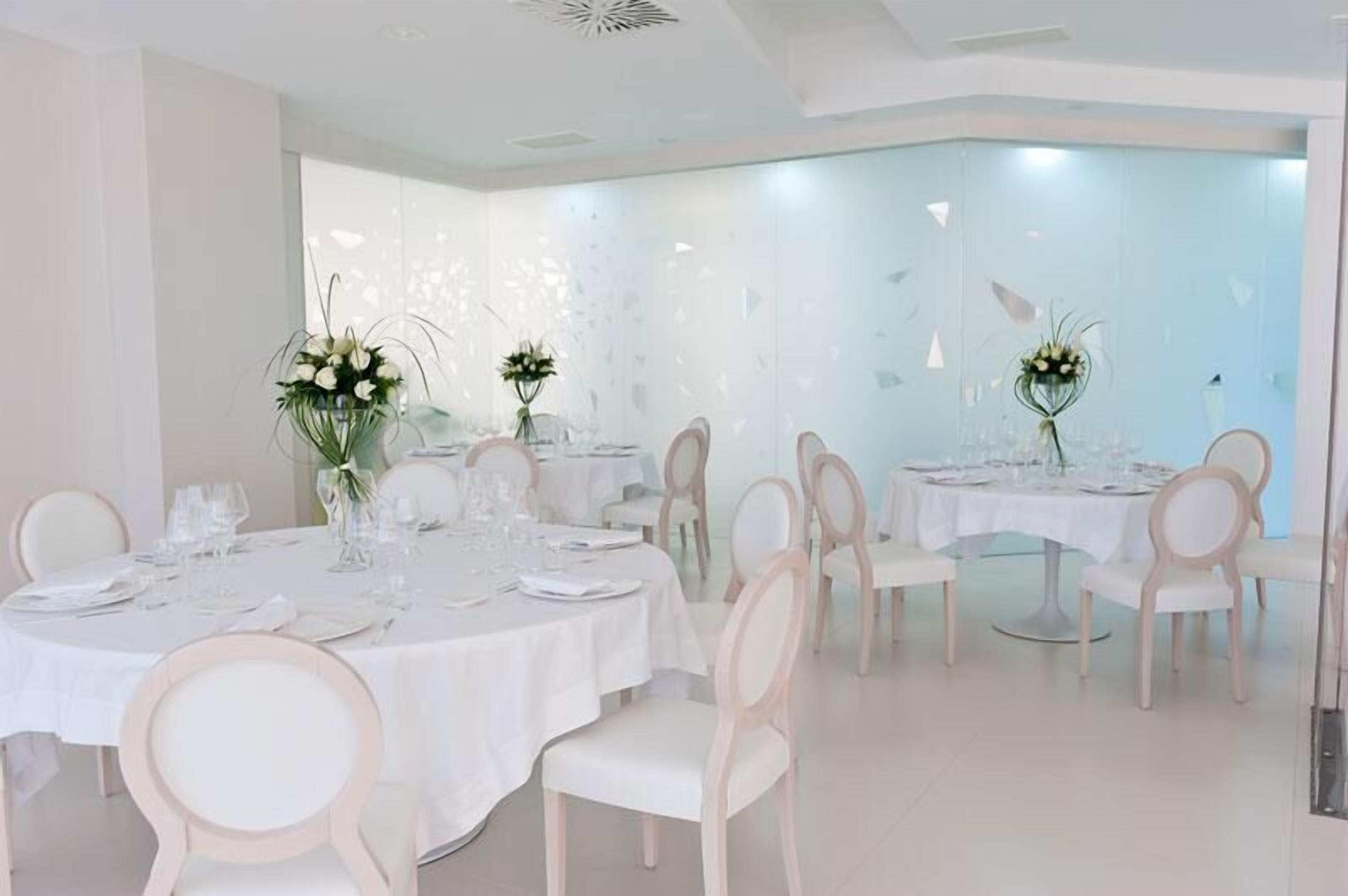 Un espace repas avec des tables rondes disposées pour accueillir quatre personnes, chacune étant recouverte d’une nappe blanche et agrémentée d’un arrangement floral.