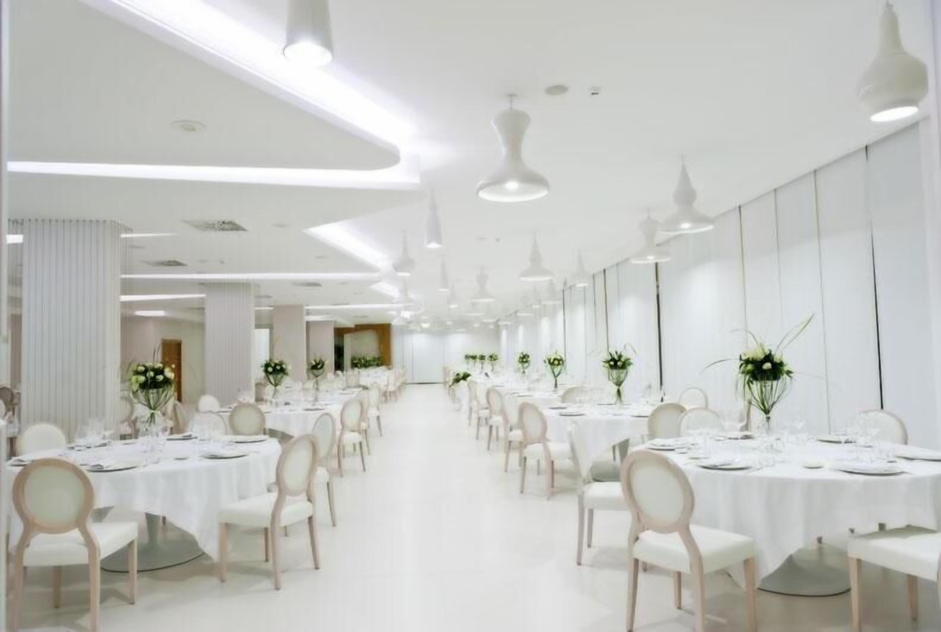 Une salle de banquet spacieuse et bien éclairée, avec de nombreuses tables rondes dressées pour le repas, des chaises blanches et des luminaires suspendus.
