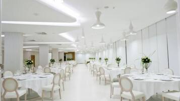 Une salle de banquet spacieuse et bien éclairée, avec de nombreuses tables rondes dressées pour le repas, des chaises blanches et des luminaires suspendus.