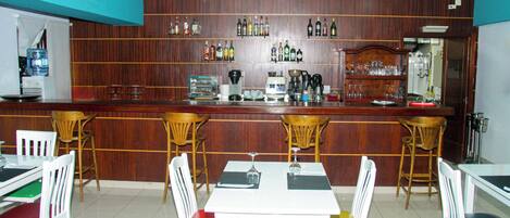 Un espace bar avec des panneaux de bois, un comptoir où sont exposées différentes bouteilles, et un coin salon avec des chaises blanches et une table.