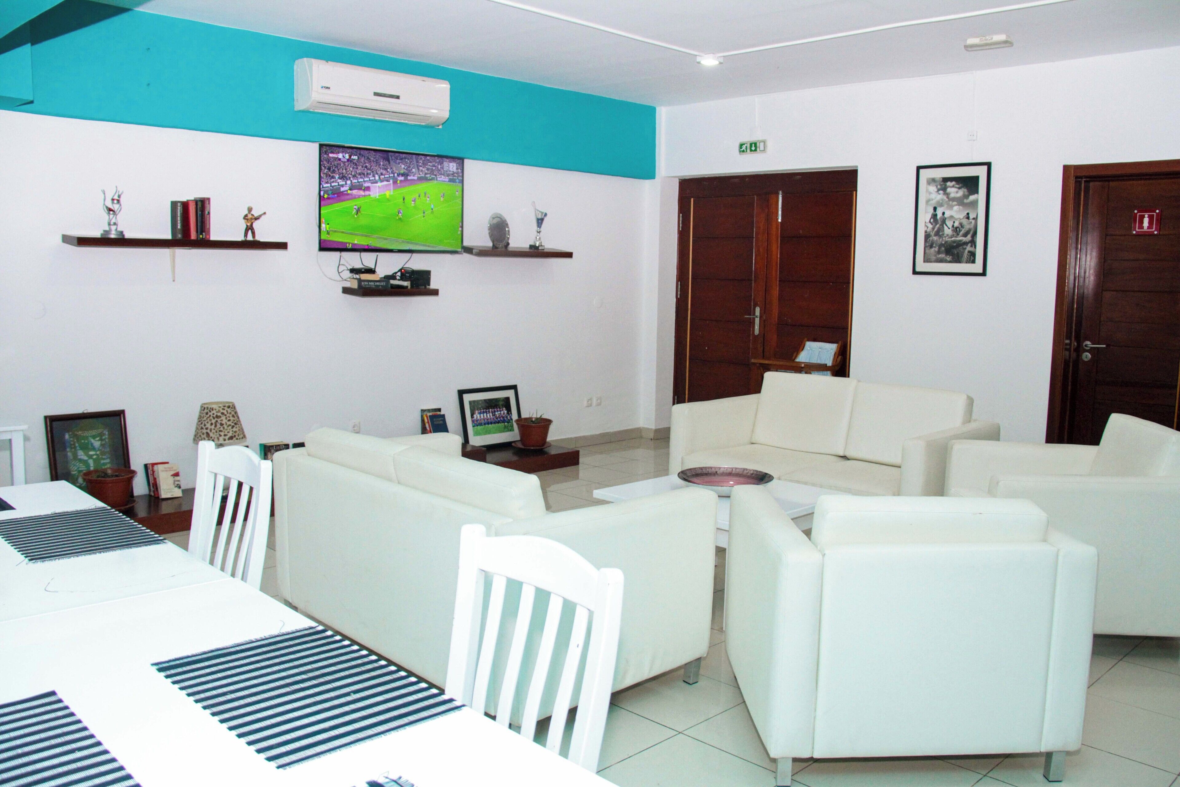 A modern living room with a white sofa set, a flat-screen TV mounted on the wall, and a dining area with a white table and chairs.