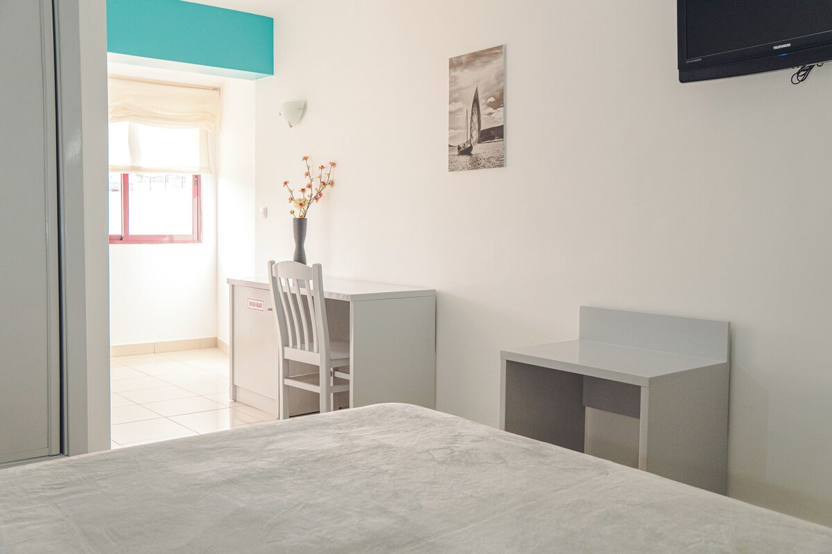 Quarto com cama, escrivaninha, cadeira, TV fixada na parede e janela com vista para o exterior.