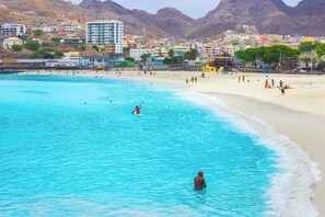 A beach with clear turquoise water, people swimming and sunbathing, and buildings in the background. - Hotel LIVVO Don Paco (São Vicente)