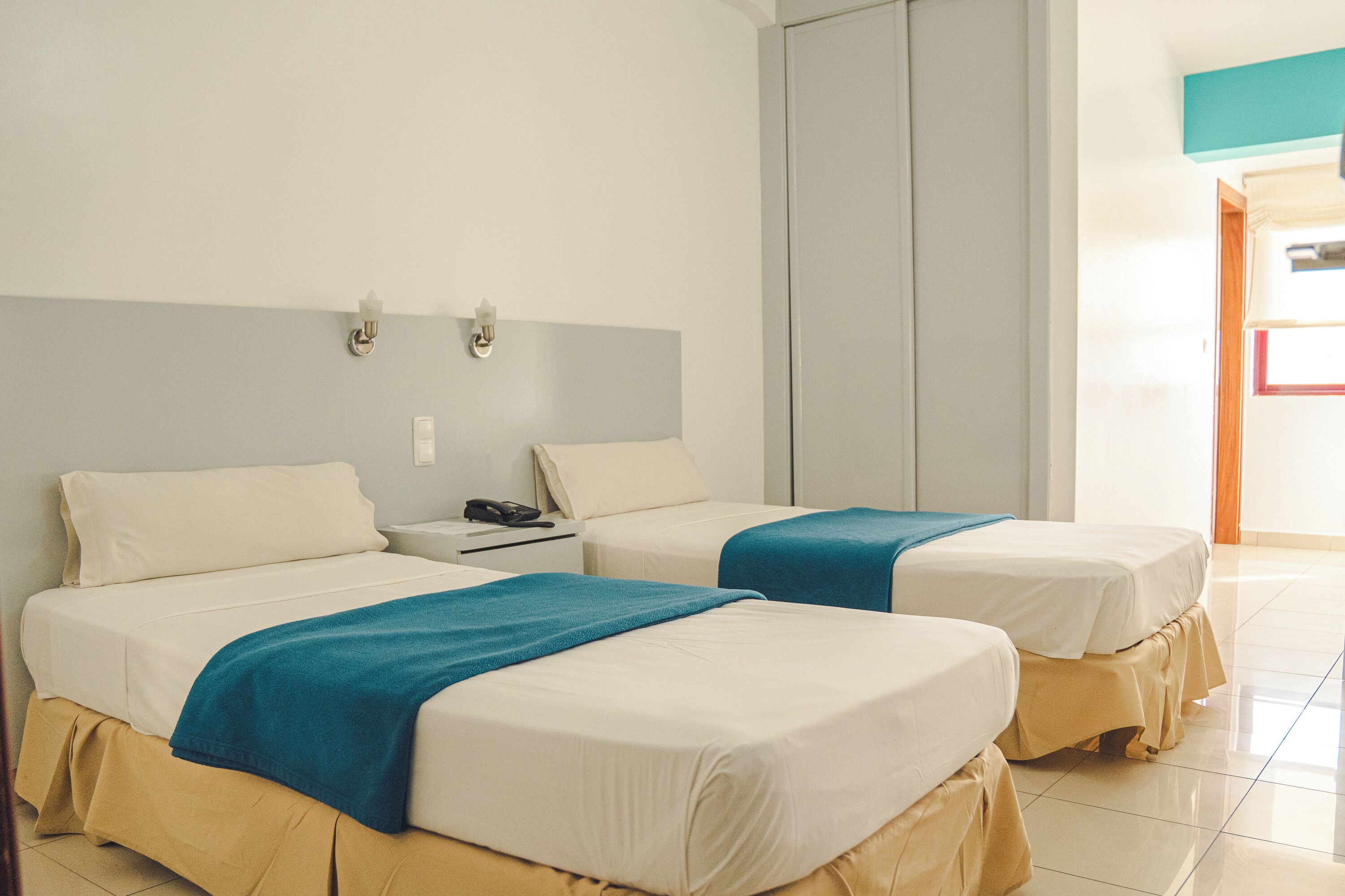 A hotel room with two beds, each with a blue blanket, white bedding, and beige curtains. There is a nightstand with a phone and a lamp between the beds.