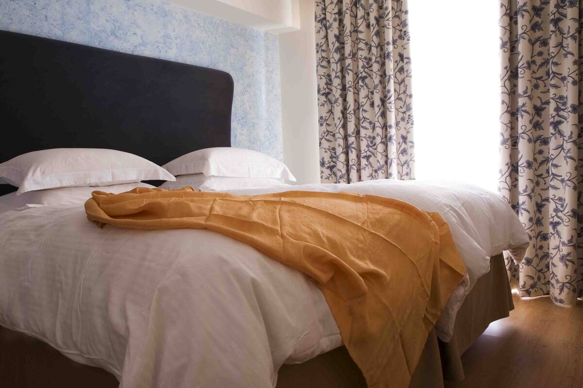 A neatly made bed with white linens and a mustard-colored throw blanket, a dark headboard, and floral-patterned curtains.