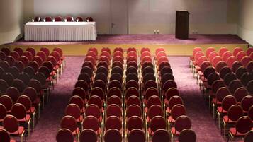 A conference room with rows of red chairs, a stage with a table and chairs, and a podium.