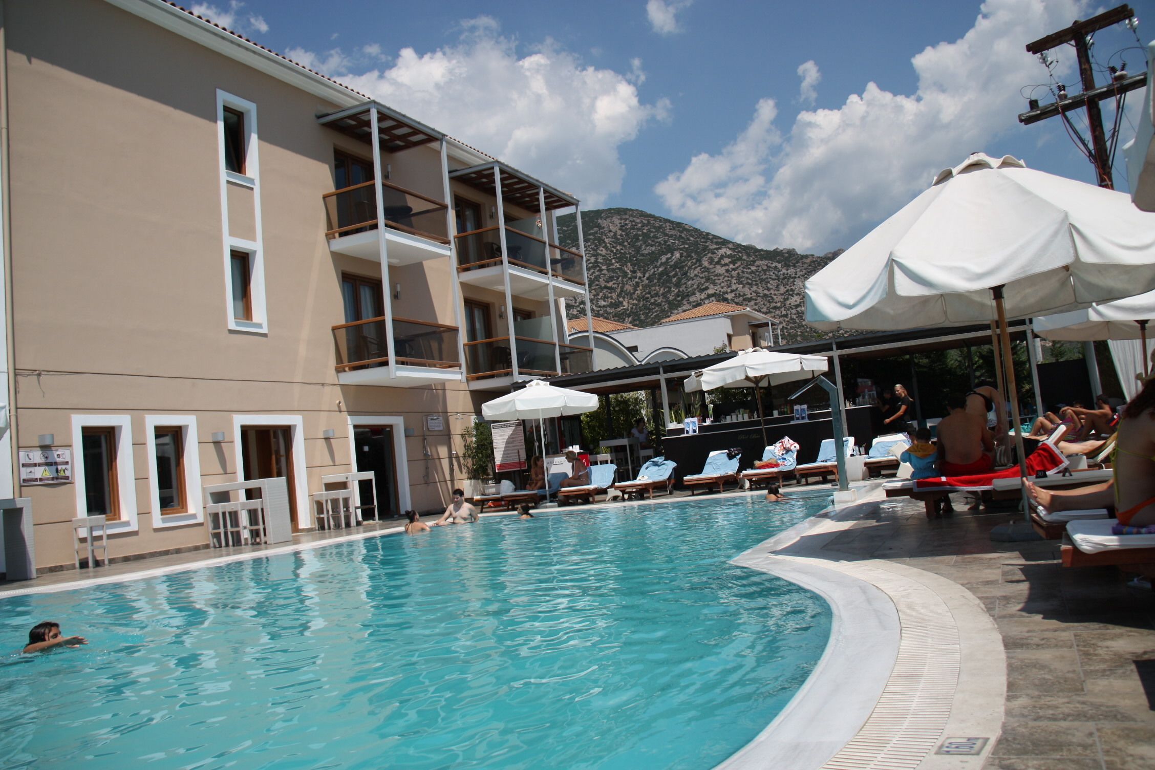 Zona de piscina con sillones de descanso y sombrillas, un edificio con balcones y montañas al fondo.