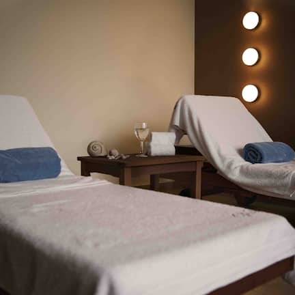 A room with two beds, each with a towel and a glass of water. There is a nightstand with a clock and a small decorative item.