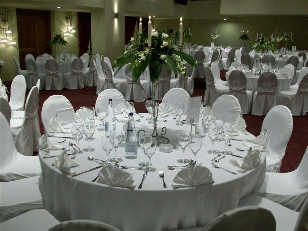 Un salón de banquetes con mesas redondas dispuestas para un evento formal, cada una con manteles blancos, cristalería y centros de mesa florales.