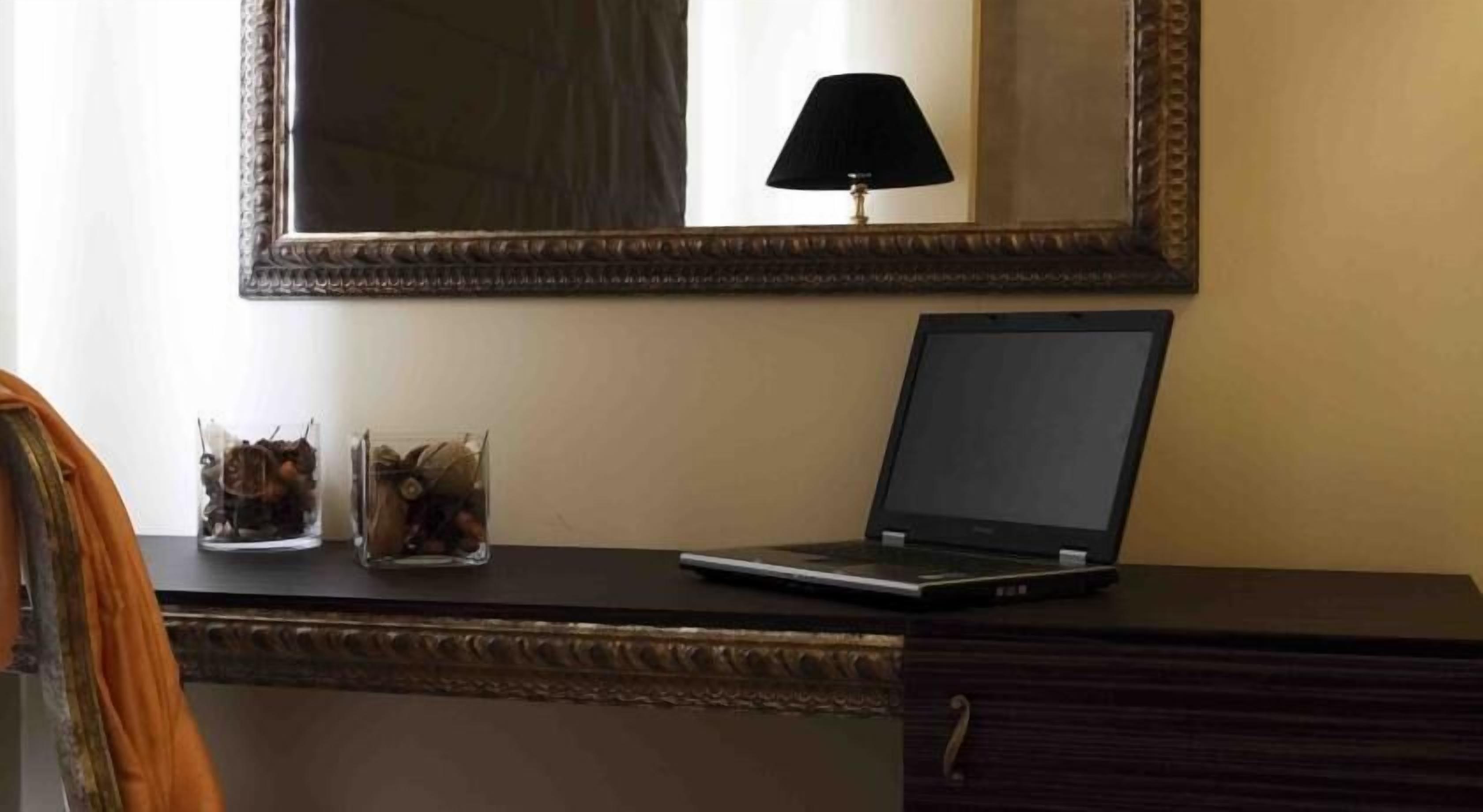 Un portatile su una scrivania con uno specchio sopra, una lampada e due contenitori di vetro con oggetti decorativi.