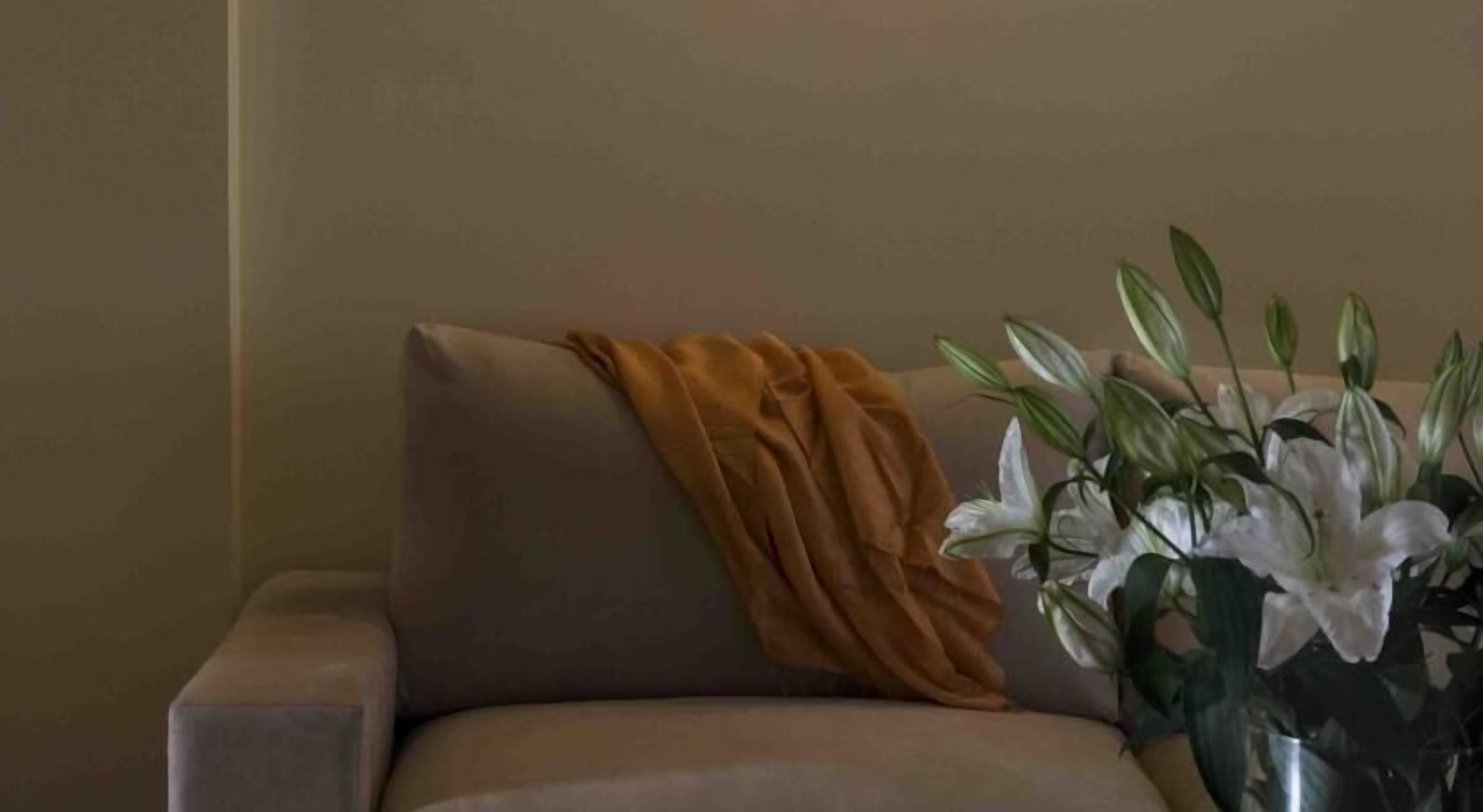 Un fauteuil beige agrémenté d’un plaid de couleur moutarde et d’un vase rempli de lys blancs.