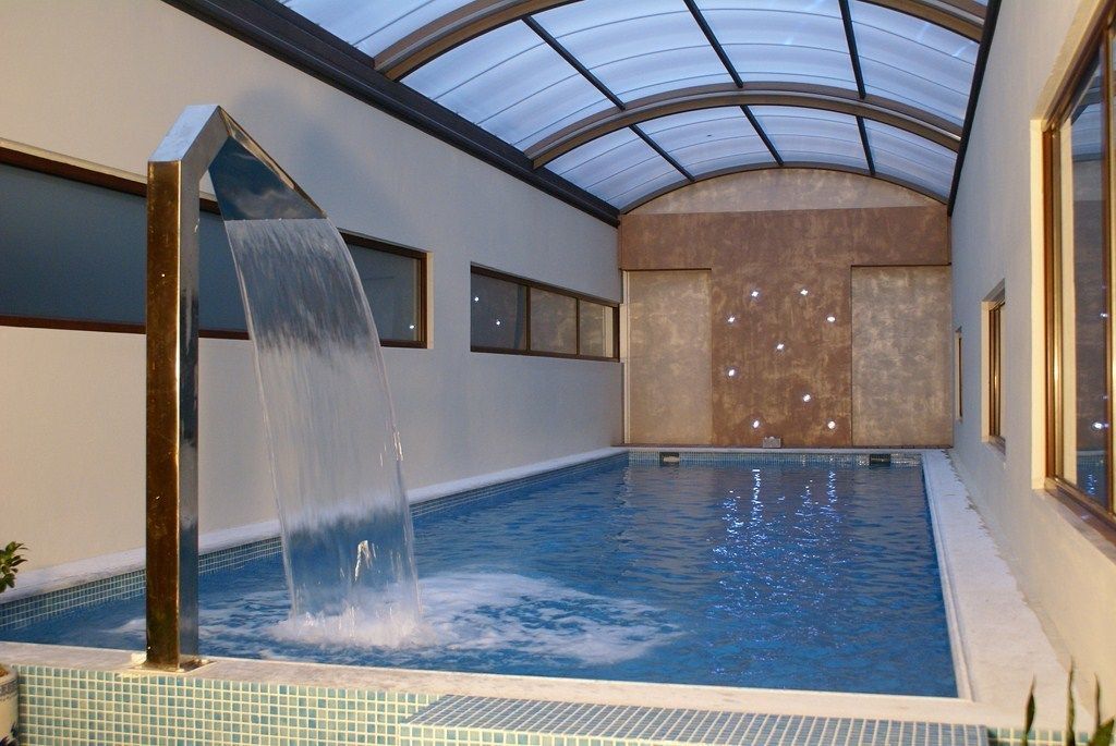 An indoor swimming pool with a waterfall feature, a glass ceiling, and a tiled floor.