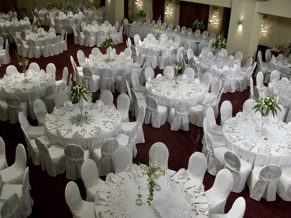 A banquet hall with multiple round tables set for dining, each with white tablecloths and chairs, and small centerpieces.