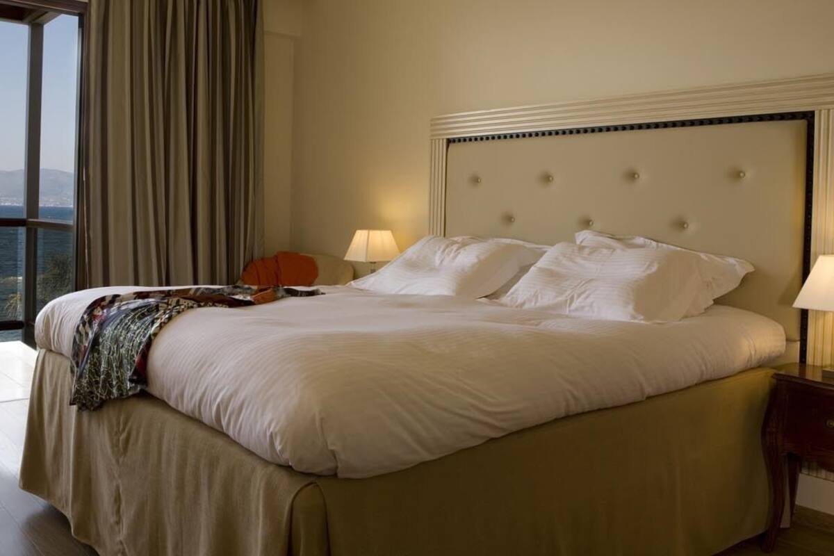 A large bed with a tufted headboard, two bedside lamps, and a view of the sea through a large window.