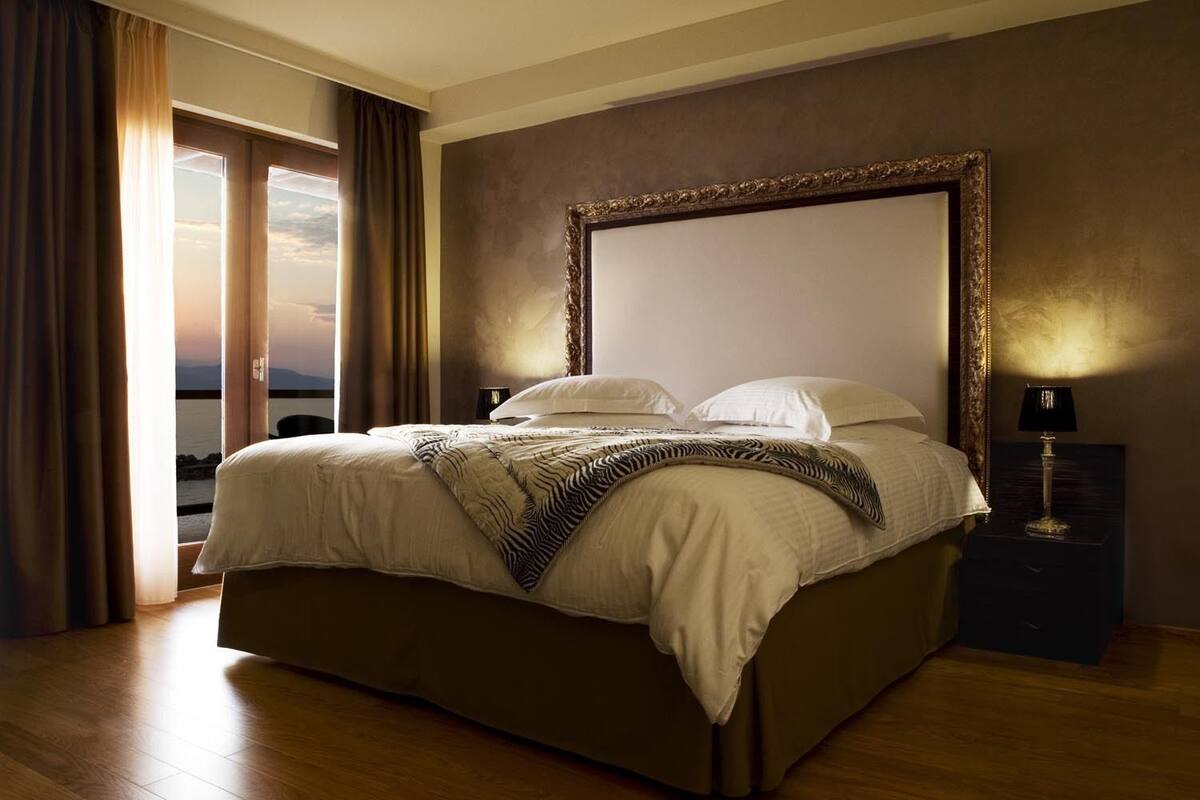 A large bed with a zebra-patterned throw, a nightstand with a lamp, and a view of the sea through a window with curtains.