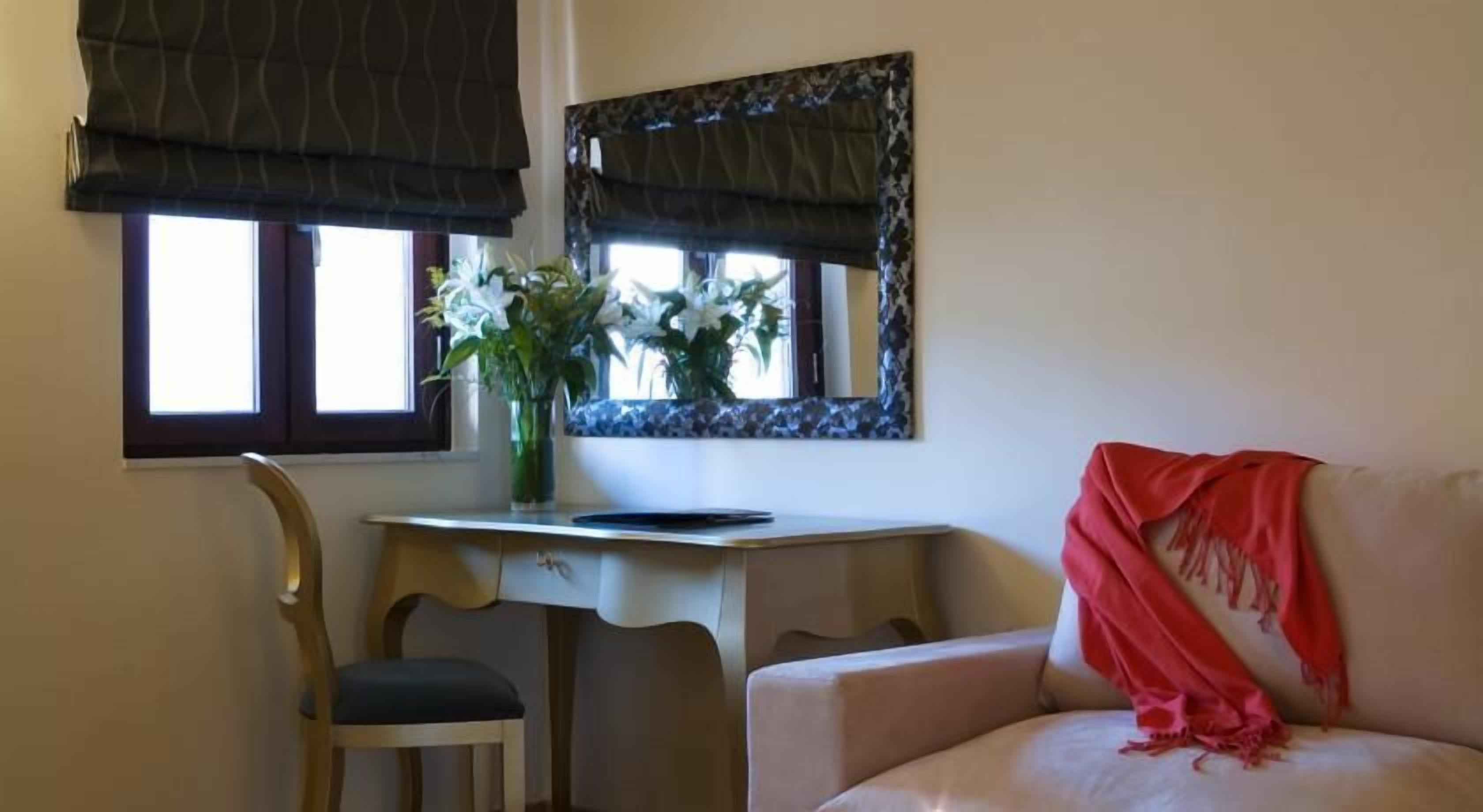 A room with a desk, chair, and a couch. There is a window with a view of the outside, and a vase with flowers on the desk.