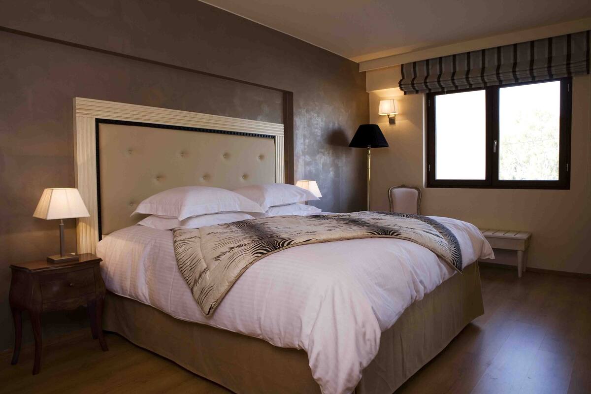 A large bed with a tufted headboard, two bedside lamps, a bench, and a window with a view.
