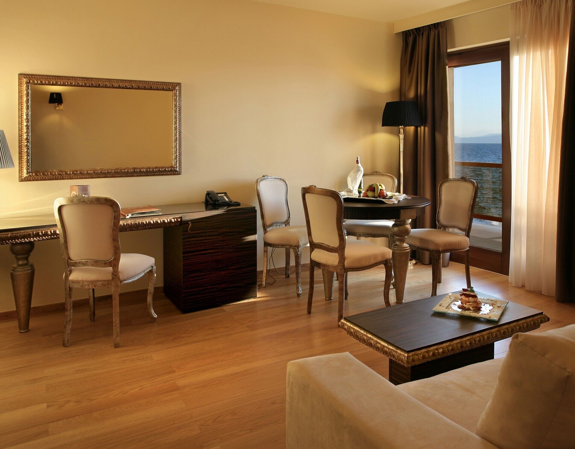 A hotel room with a wooden desk, two chairs, a round table with a lamp, a large mirror, and a view of the sea through a window.