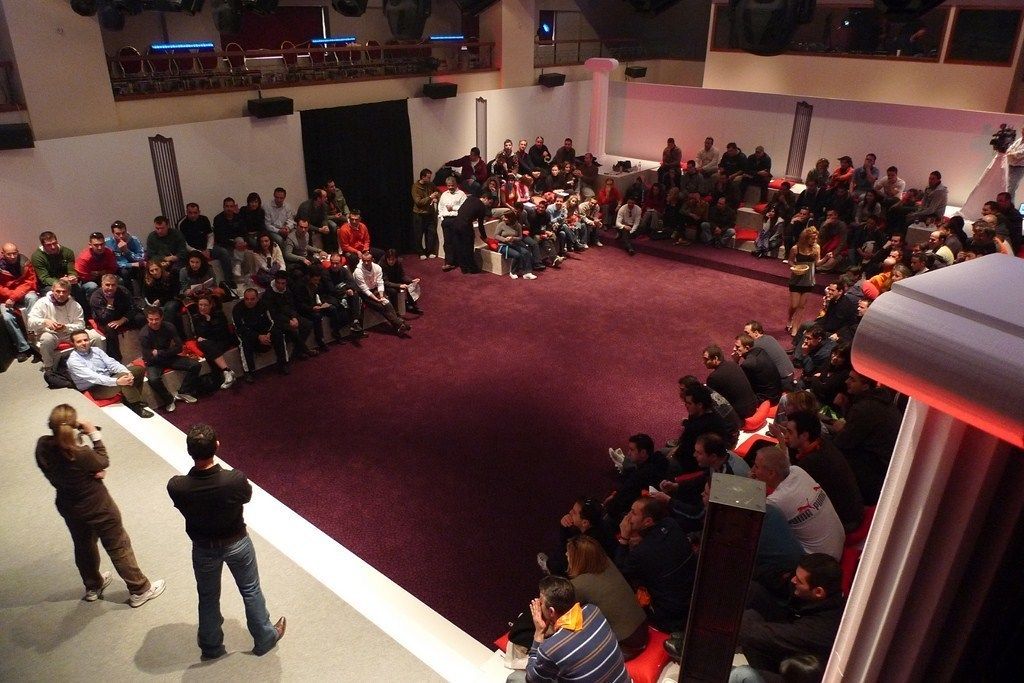 Un amplio espacio interior con piso alfombrado en rojo, asientos escalonados y un escenario al frente.