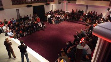 Un amplio espacio interior con piso alfombrado en rojo, asientos escalonados y un escenario al frente.