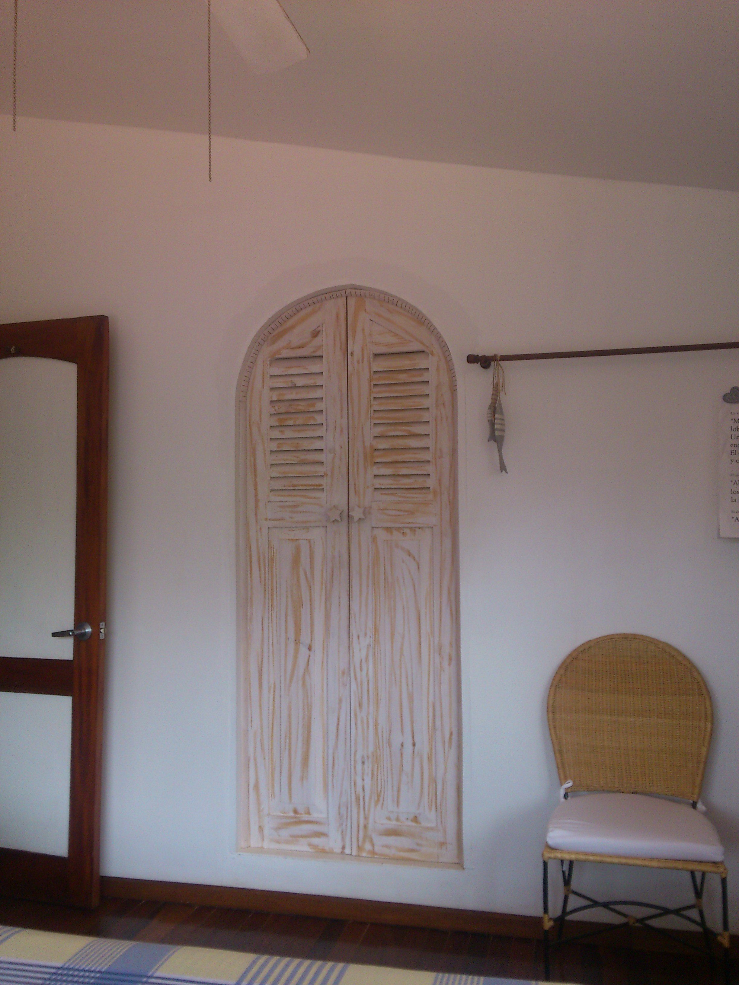 Una habitación con puerta de madera, un amplio ventanal de arco con contraventanas, una silla de mimbre y un aplique de pared.